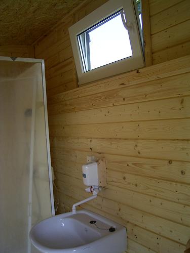 Wnętrze sanitariatu w drewnianej konstrukcji z uchylnym oknem, umywalką i dozownikiem mydła na ścianie z desek.